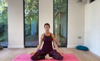 Marcia Mercer in a seated yoga pose on a pink mat in a white walled yoga studio with tall, floor to ceiling windows showing a courtyard garden