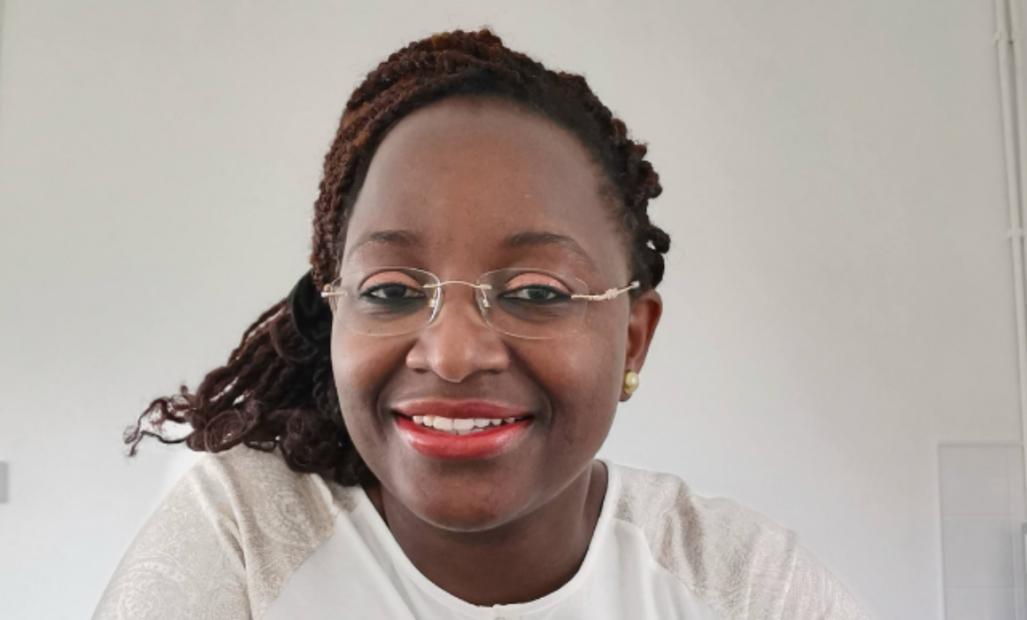 A smiling Bami Adenipekun looking at the camera, wearing a white top, glasses and red lipstick
