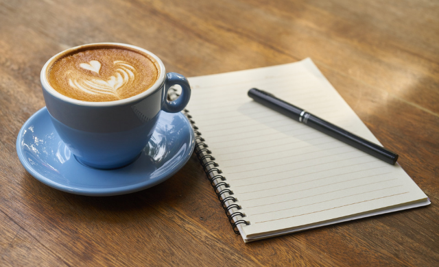 Mid blue coloured coffee cup and saucer on a wood table next to a blank ring bound note pad and pen