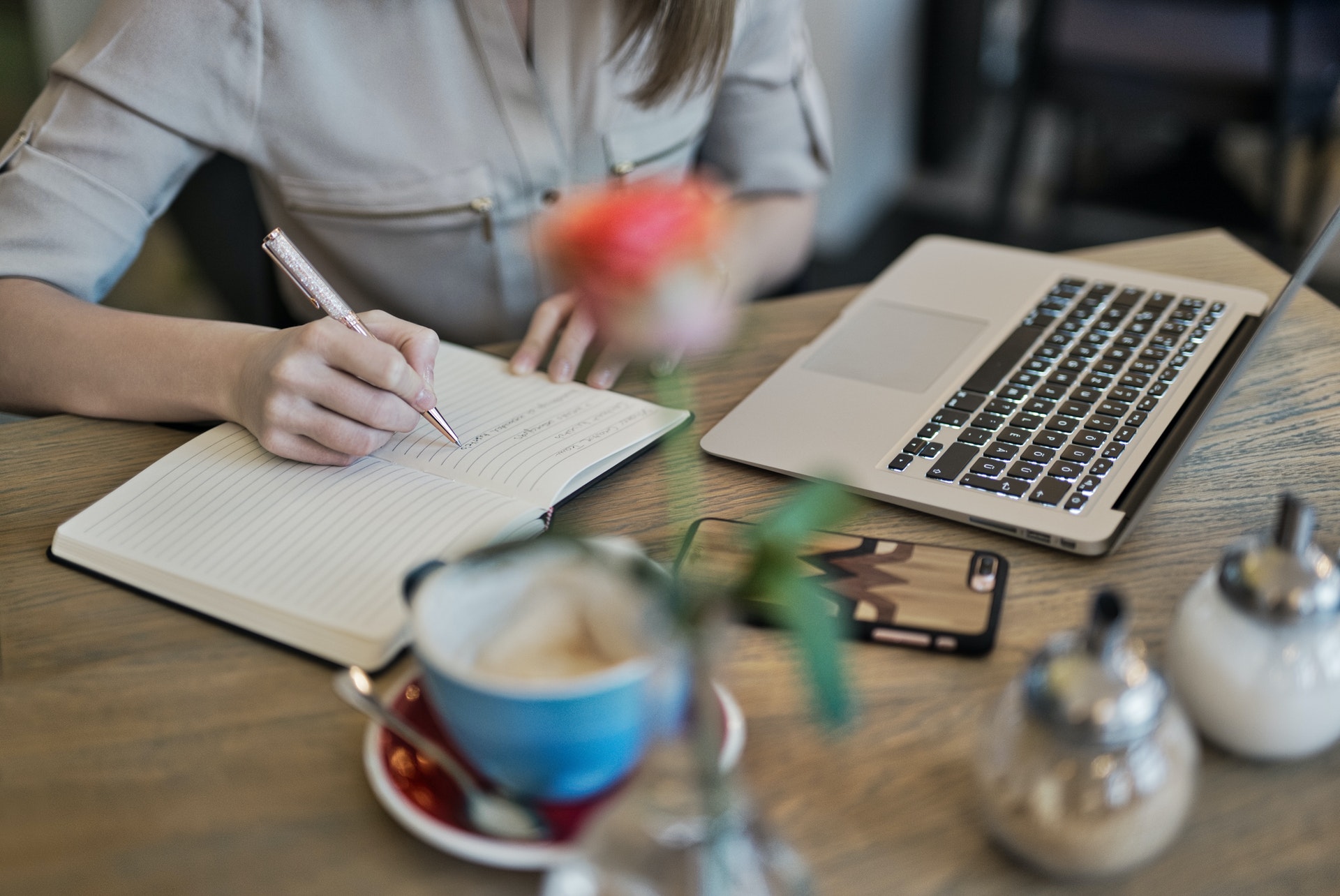 Woman sat at a table, writing in a notebook, coffee cup, laptop, salt & pepper shaker and a vase with a single orange rose slightly out of focus